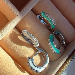 May include: Three pairs of silver hoop earrings with white and turquoise enamel. The earrings have a small hinge and are decorated with clear rhinestones.