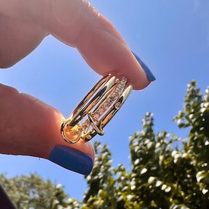 May include: A gold ring with three bands, each set with small clear stones. The ring is held up against a blue sky.