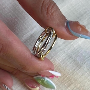 May include: A two-tone ring with gold and silver bands, featuring a row of small, sparkling stones. The ring has an intricate design with interlocking elements. The ring is held between fingers, showcasing its elegant details.