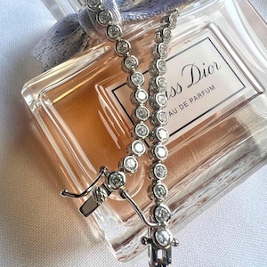 May include: A silver-toned tennis bracelet featuring a continuous row of round, clear stones in bezel settings, complete with a secure box clasp. The bracelet is draped over a rectangular glass perfume bottle labeled Miss Dior Eau de Parfum.