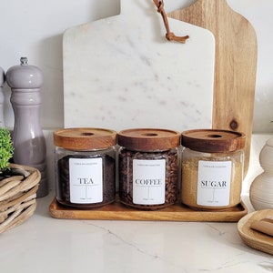 May include: Three glass jars with wooden lids and labels that read "Tea", "Coffee", and "Sugar". The jars are sitting on a wooden shelf with two chopping boards behind them. One chopping board is white marble and the other is brown wood.