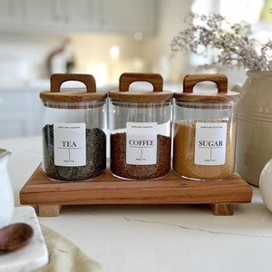 Op de afbeelding: Drie glazen potten met houten deksels en etiketten met de tekst "Thee", "Koffie" en "Suiker". De potten staan op een houten dienblad.