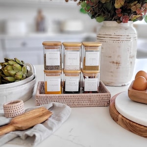 May include: Six glass spice jars with bamboo lids and white labels. The labels have the names of the spices: oregano, sea salt, cumin, turmeric, black peppercorns, and cinnamon sticks. The jars are arranged in two rows of three on a pink wicker tray.