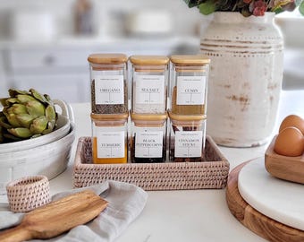 May include: Six glass spice jars with bamboo lids and white labels. The labels have the names of the spices: oregano, sea salt, cumin, turmeric, black peppercorns, and cinnamon sticks. The jars are arranged in two rows of three on a pink wicker tray.