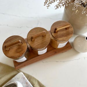 Puede incluir: Tres tarros de vidrio con tapas y asas de madera, llenos de varios granos, en una bandeja de madera. Los tarros est&aacute;n etiquetados con etiquetas blancas.