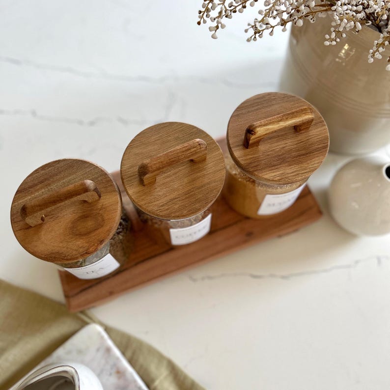 Puede incluir: Tres tarros de vidrio con tapas y asas de madera, llenos de varios granos, en una bandeja de madera. Los tarros est&aacute;n etiquetados con etiquetas blancas.