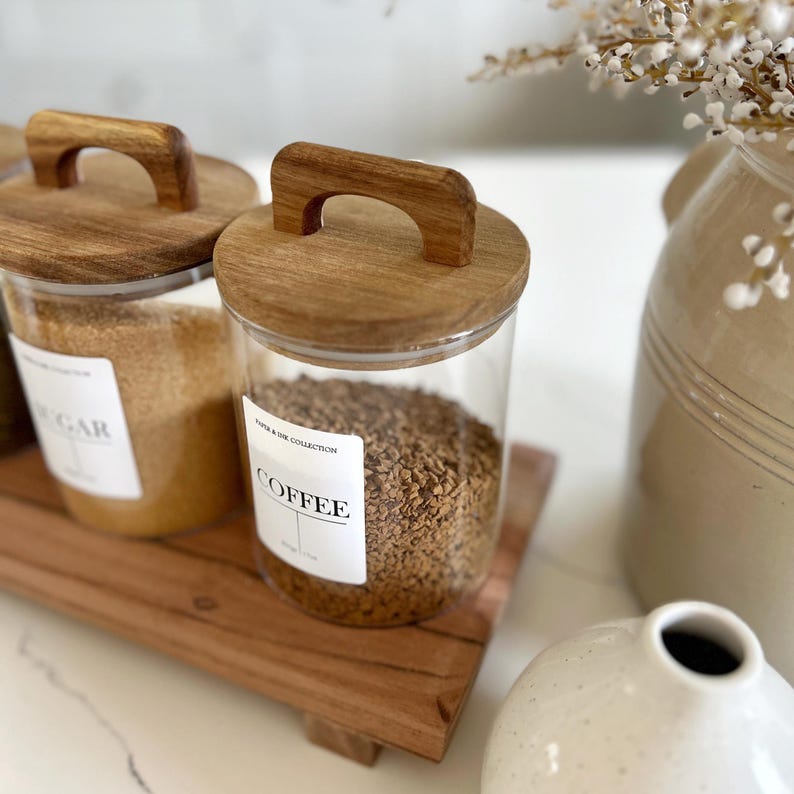 Puede incluir: Tres botes de vidrio con tapas y asas de madera, colocados sobre una bandeja de madera. Los botes est&aacute;n llenos de az&uacute;car, caf&eacute; y t&eacute;. Las etiquetas en los botes dicen "Az&uacute;car", "Caf&eacute;" y "T&eacute;".