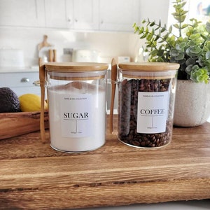 Op de afbeelding: Twee glazen potten met bamboe deksels en houten lepels. De potten zijn gevuld met suiker en koffiebonen. De potten zijn gelabeld met "Suiker" en "Koffie".