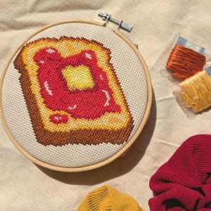 Puede incluir: Un bastidor de bordado de punto de cruz con una rebanada de pan tostado con mantequilla y mermelada. La tostada es marrón, la mantequilla es amarilla y la mermelada es roja. Hilos de bordar en naranja y amarillo están junto al bastidor. Tela roja y amarilla en primer plano.