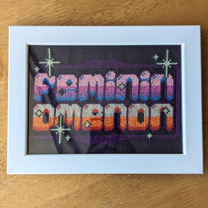 May include: A framed cross-stitch piece with the words "Feminine Demon" in a pixelated font. The letters are in shades of pink, purple, blue, orange, and yellow. The background is black with white stars.