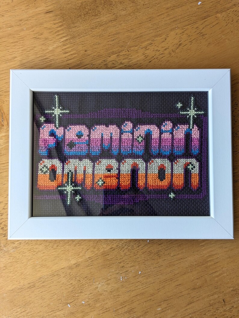 May include: A framed cross-stitch piece with the words "Feminine Demon" in a pixelated font. The letters are in shades of pink, purple, blue, orange, and yellow. The background is black with white stars.