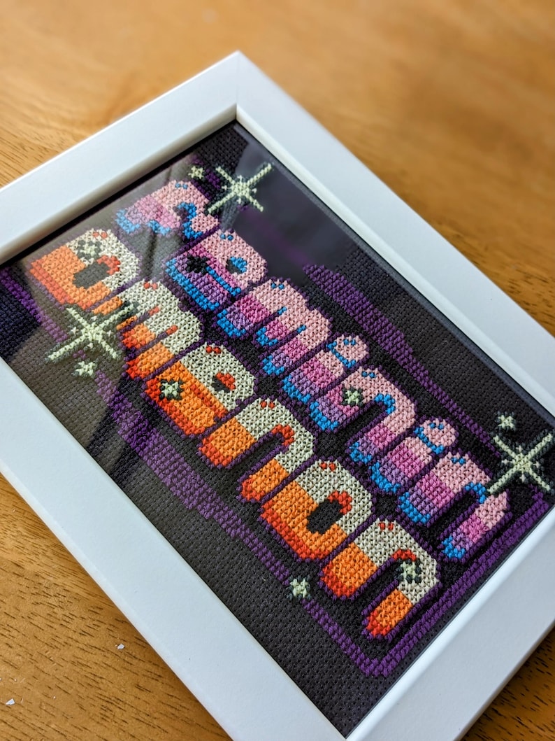 May include: A framed cross-stitch with the words "Remarkable Human" in a colorful, pixelated font. The background is a dark purple with white stars.