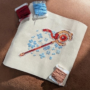 Puede incluir: Bordado de punto de cruz sobre tela beige con un diseño de bastón rojo y dorado con detalles florales azules. Tres carretes de hilo en rojo, azul y marrón sugieren un proyecto de manualidades.
