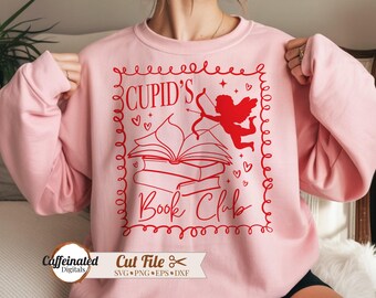May include: A light pink sweatshirt with a red graphic that reads "Cupid's Book Club." The design features Cupid with a bow and arrow, books, hearts, and stars. The sweatshirt is a long-sleeved crew neck.