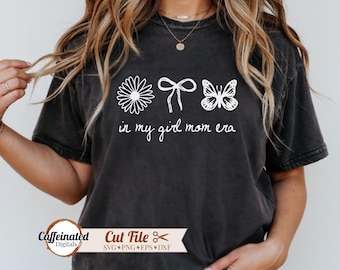 May include: Dark gray t-shirt with a white daisy, bow, and butterfly design, along with the text "in my girl mom era." The shirt also features the text "Caffeinated Digitals" and "Cut File" with file type icons.