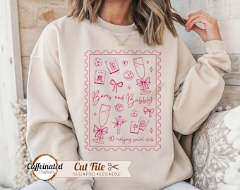 May include: Beige sweatshirt with a pink graphic design featuring champagne glasses, mahjong tiles, and the text "Bams and Bubbly Mahjong Social Club." The design is framed within a pink postage stamp border.