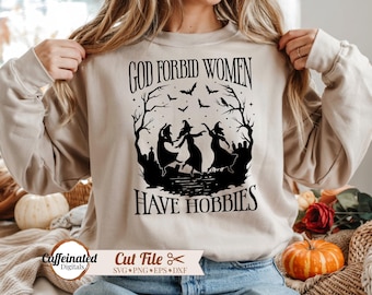 May include: A beige long-sleeve sweatshirt featuring a black graphic print of three witches dancing in a graveyard under a night sky with bats and bare trees. The text "GOD FORBID WOMEN HAVE HOBBIES" is printed in a bold, stylized black font.