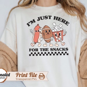 May include: White sweatshirt with the text "I'm just here for the snacks" in black.  A cartoon hot dog, football, and popcorn bucket are walking together.