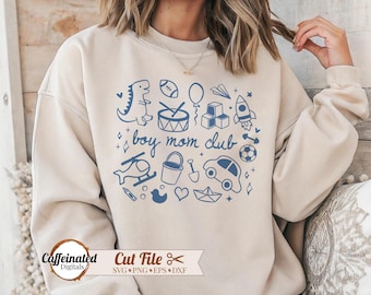 May include: A beige sweatshirt with a blue graphic design featuring various toy illustrations and the text "boy mom club." The design includes a dinosaur, football, drum, rocket, car, and other playful elements.