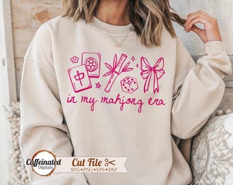 May include: A beige sweatshirt with a pink graphic design featuring Mahjong tiles, bamboo, a dice, and a bow. The text "in my mahjong era" is written in a cursive font. The bottom left corner has the text "Caffeinated Digitals".