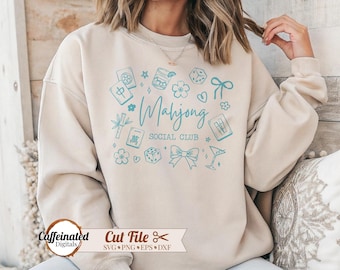 May include: Beige sweatshirt with a teal graphic that reads "Mahjong Social Club." The design includes various Mahjong-related icons such as tiles, dice, and flowers. The sweatshirt has a crew neck and long sleeves.