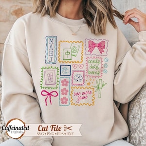May include: A beige sweatshirt featuring colorful embroidered patches with Mahjong-themed graphics. The patches include text, dice, flowers, and a bow. The phrase "one more game" is also present. The sweatshirt has a relaxed fit.