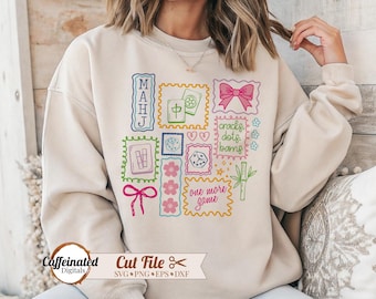 May include: A beige sweatshirt featuring colorful embroidered patches with Mahjong-themed graphics. The patches include text, dice, flowers, and a bow. The phrase "one more game" is also present. The sweatshirt has a relaxed fit.