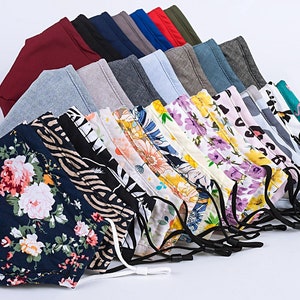 May include: A stack of 15 colorful fabric face masks with various patterns, including floral, geometric, and abstract designs. The masks are arranged in a fan shape, with the bottom layer showing a dark blue mask with a floral pattern.