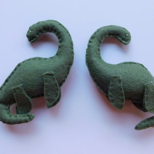 Può includere: Due giocattoli di dinosauri in feltro verde fatti a mano. Ogni dinosauro ha un collo lungo, piccole zampe e una coda curva. Il feltro è cucito con un filo a contrasto. I dinosauri sono posizionati su una superficie bianca.