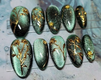 May include: Ten green and gold oval-shaped false nails with a floral design. The nails have a shimmery, iridescent finish.