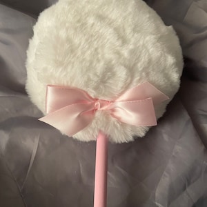 May include: A white fluffy powder puff with a pink satin bow attached to a pink wooden stick.