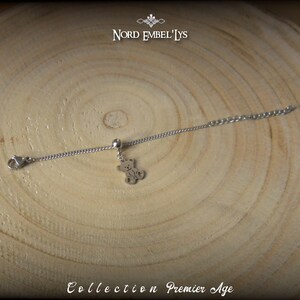 Puede incluir: Un brazalete de cadena de plata con un pequeño dije de oso. El brazalete está sobre una superficie de madera. Collection Premier Age.