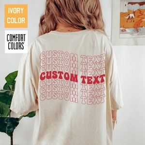 May include: Ivory t-shirt featuring the words "CUSTOM TEXT" in red, repeated vertically on the back. The shirt is a Comfort Colors brand. The text is in a retro, wavy font.