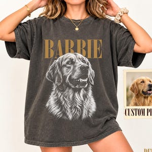 Op de afbeelding: Een antracietgrijs t-shirt met korte mouwen voorzien van een grote zwart-witillustratie van een golden retriever met de tekst BARBIE in gouden letters erboven. Een klein vierkant inzetstuk toont een kleurenfoto van de hond met de tekst CUSTOM PHOTO.
