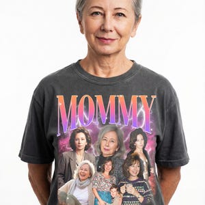 May include: A woman wearing a dark gray t-shirt with the word "MOMMY" in a gradient pink and orange font. The shirt features a collage of images of women. The woman in the image has gray hair and is smiling.