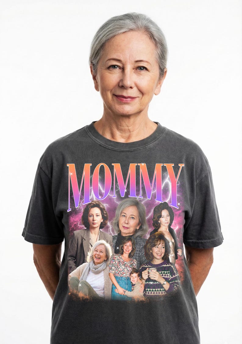 May include: A woman wearing a dark gray t-shirt with the word "MOMMY" in a gradient pink and orange font. The shirt features a collage of images of women. The woman in the image has gray hair and is smiling.