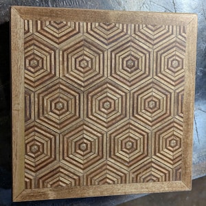 May include: Square wooden tray with a geometric pattern of interlocking hexagons. The tray features a light brown frame and a mosaic-like design in varying shades of brown and tan. The design is symmetrical and detailed.