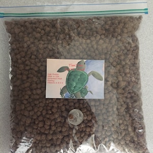May include: A clear plastic bag filled with brown turtle food pellets. The bag has a label that says "Tim-Tam Turtle Food" and lists the ingredients as "High Protein, Low Fat, Guaranteed Fresh, Enhanced with Vitamins A, B, D, D3, E".