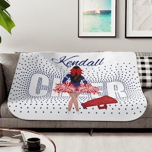 May include: White fleece blanket with a blue and red cheerleader design. The blanket features a girl in a cheerleading uniform holding pom-poms and a megaphone. The text "Kendall Cheer" is written on the blanket.