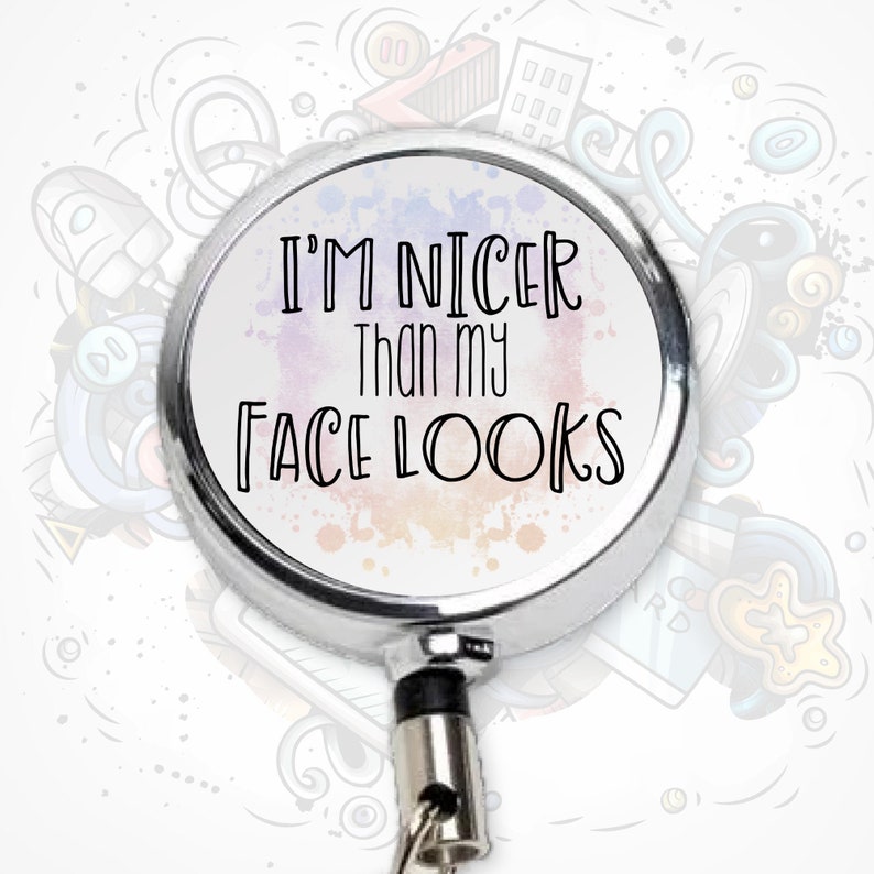 May include: A silver retractable badge holder with a white background and colorful watercolor splatter design. The text "I'm nicer than my face looks" is printed in black.