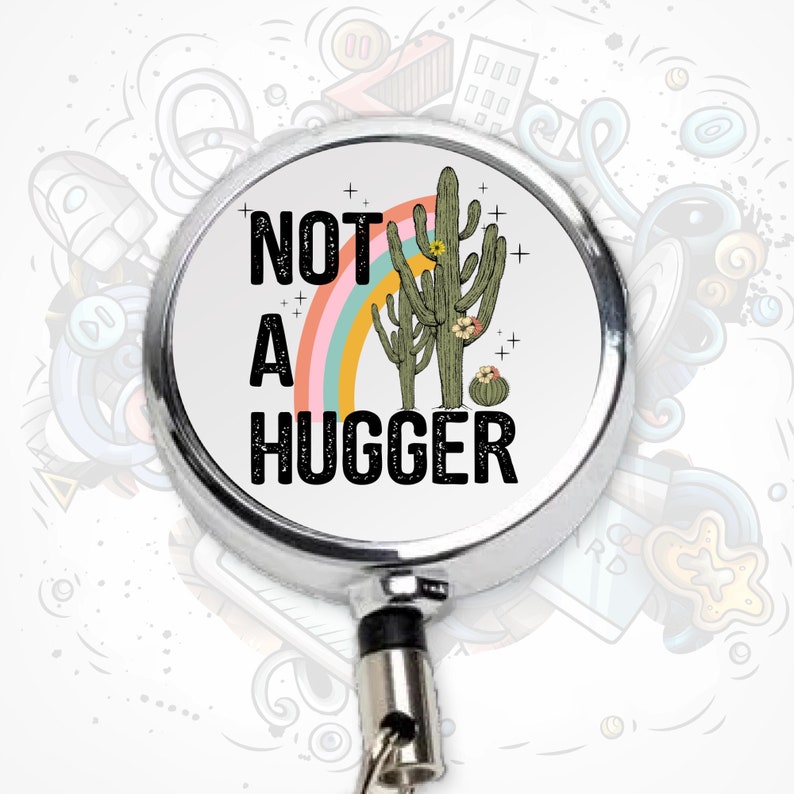 Puede incluir: Un portagafete retr&aacute;ctil plateado con fondo blanco y un gr&aacute;fico en blanco y negro de dos cactus con un arco&iacute;ris detr&aacute;s. El texto "NOT A HUGGER" est&aacute; impreso en negro.