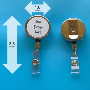 May include: A silver retractable badge holder with a clear plastic strap and a round, white, customizable area that says "Your Design Here". The holder measures 1.6 inches in diameter and 3.8 inches in height.