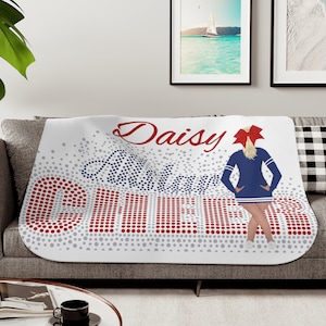 May include: White fleece blanket with a red, white, and blue cheerleading design. The blanket features a cheerleader with a red bow, the words "Daisy" and "Cheer" in a sparkly font, and a background of red and gray dots.