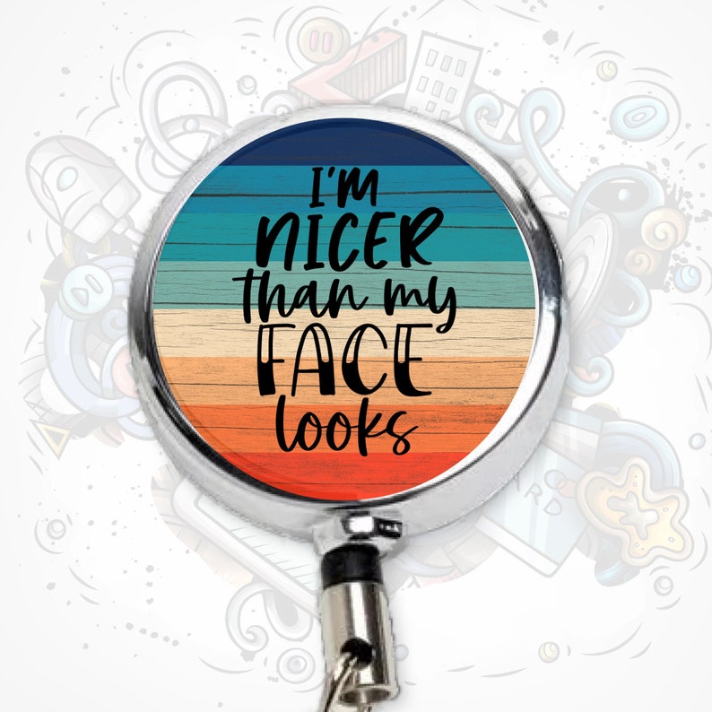 May include: A silver retractable badge reel with a round, colorful design. The design features a woodgrain pattern in shades of blue, yellow, and orange. The text "I'm nicer than my face looks" is printed in black on the design.