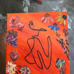 Pode incluir: Pintura em tela quadrada com fundo laranja, apresentando uma figura de arte de linha preta e detalhes florais. As flores incluem rosas cor-de-rosa, margaridas azuis e amarelas e cachos roxos. A obra de arte está em uma tela quadrada.