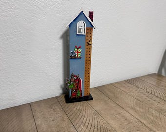 May include: A blue wooden Christmas countdown house with a Santa Claus figure climbing a ladder. The house has a window and a ruler with numbers 1-24 on the side. There is a star on top of the house.