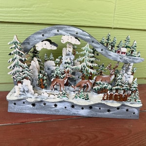 May include: A multi-layered wooden winter scene decoration featuring three brown deer standing among snow-dusted evergreen trees and rocky formations. An arched gray wooden frame with star cutouts spans the top, framing the forest landscape.
