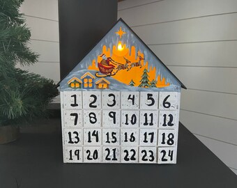 Peut inclure: Un calendrier de l'Avent de Noël en bois en forme de maison. Le toit est peint avec une scène hivernale du Père Noël dans son traîneau. Il comporte 24 tiroirs numérotés, chacun avec un numéro noir sur fond blanc.