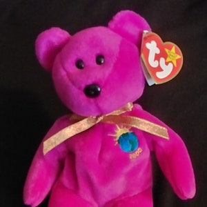 Puede incluir: Un oso de peluche rosa Ty Beanie Baby con una cinta dorada alrededor del cuello. El oso tiene un globo terráqueo azul y verde en el pecho con el año 2000 impreso en él. El oso tiene una etiqueta Ty roja unida a su oreja.