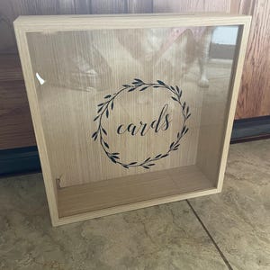 May include: A wooden box with a glass front and a black wreath with the word "cards" inside. The box is light brown and has a natural wood grain.